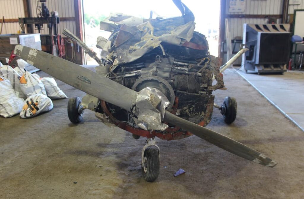 Front view of a heavily damaged aircraft engine and propeller housed in a hangar, showing bent blades and a crushed engine cowling; signs of mud and debris are present.